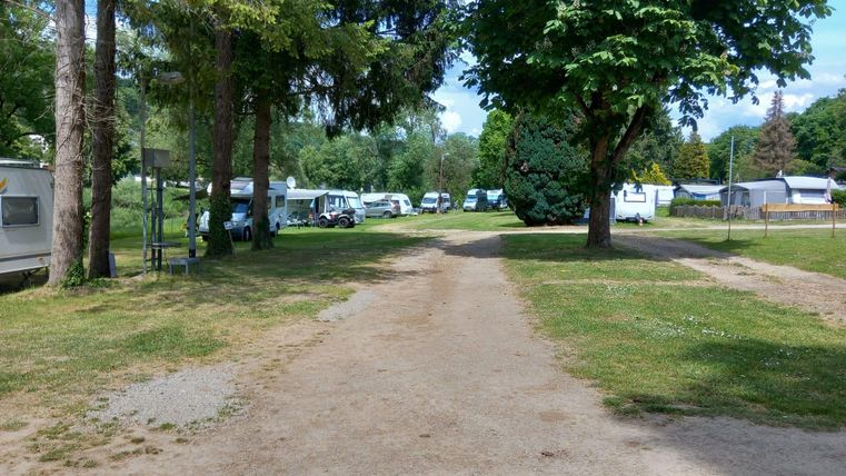 Eine ruhige Campingplatzansicht mit Wohnwagen und Bäumen. Der Weg ist von grünem Gras umgeben und führt durch die Anlage.
