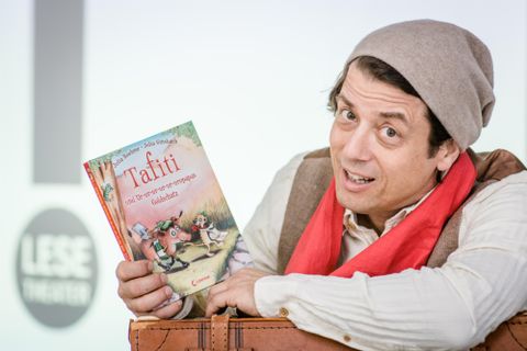 Un homme avec un chapeau gris et une écharpe rouge tient un livre pour enfants intitulé "Tafiti". En arrière-plan, on peut voir des éléments d'un événement de lecture.