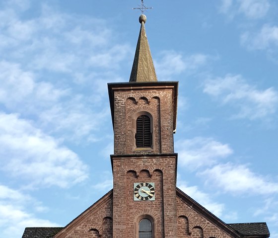Backsteinkirche St. Laurentius mit Turm und Uhr, umgeben von Bäumen und einem Kreuz im Vordergrund, unter blauem Himmel., © TI Bitburger Land