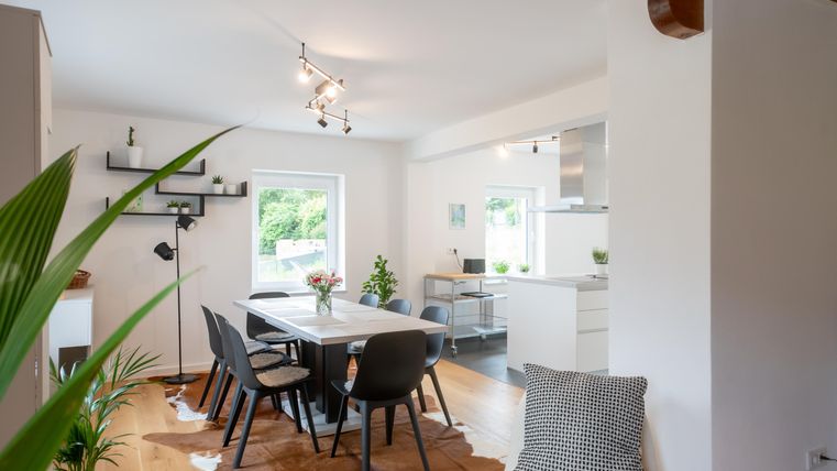 Een moderne woonkamer met een eettafel en zwarte stoelen. Grote ramen laten veel daglicht binnen en planten zorgen voor een gezellige sfeer.