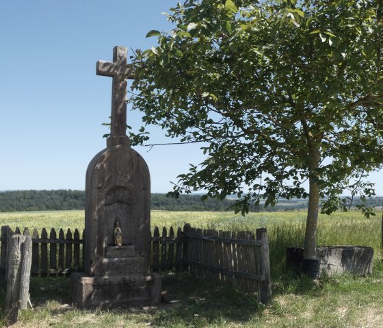 Steinernes Wegekreuz neben einem Baum auf einem Feldweg, umgeben von einem Holzzaun. Im Hintergrund Felder und Wald., © TI Bitburger Land