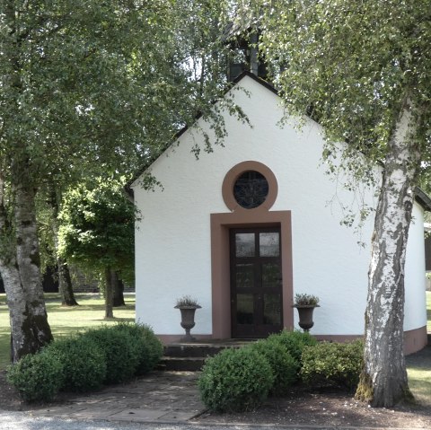 Une petite chapelle blanche aux accents bruns, entourée d'arbres et d'arbustes dans un paysage verdoyant., © TI Bitburger Land