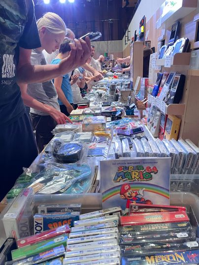 Een drukke vlooienmarkt met tafels vol videogames en consoles. Veel kopers doorzoeken de aangeboden producten.