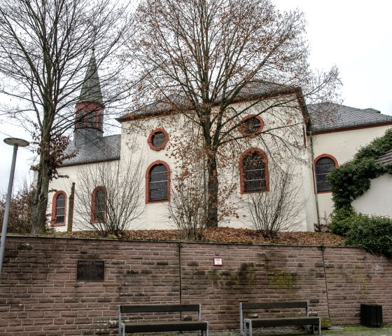 Eine Kirche in Wißmannsdorf mit roten Fensterrahmen, umgeben von kahlen Bäumen und einer Steinmauer im Vordergrund., © Tourist-Info Bitburger Land