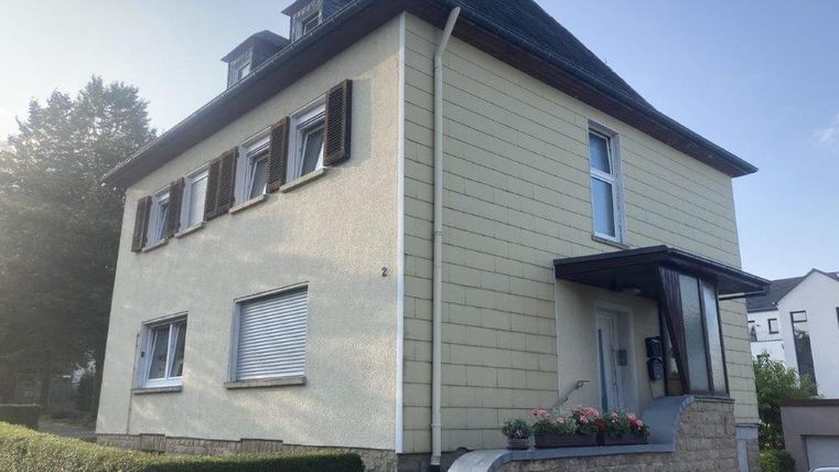 Ein modernes, zweigeschossiges Wohnhaus mit hellem Außenputz und schwarzen Fensterläden. Vor dem Haus befindet sich eine kleine Bepflanzung und ein gepflasterter Bereich.