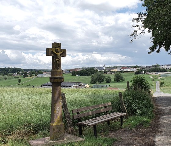 Een wegkruis en een bankje staan aan de rand van het pad met uitzicht op de wijk Mötsch, omgeven door groene velden en een bewolkte hemel.