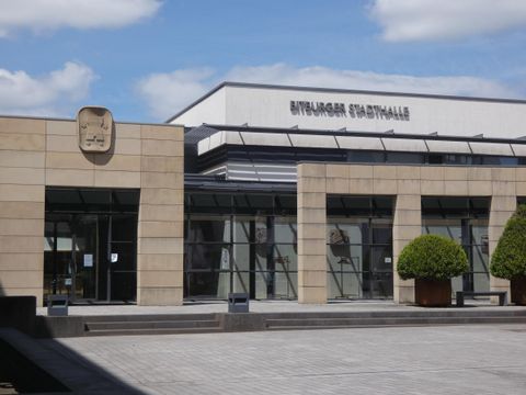 The town hall in Einburg is a modern building with a clear glass facade. In the foreground, there are decorative plants and a paved square.
