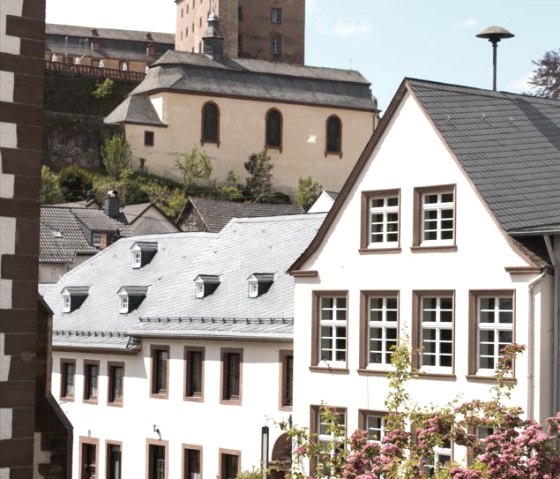Malberg Castle towers in the background, surrounded by modern buildings and flowering bushes in the foreground. The sky is slightly cloudy., © TI Bitburger Land