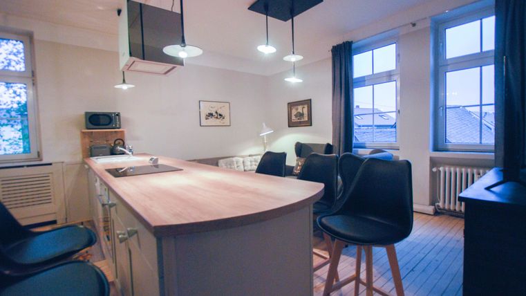 Eine moderne, helle Küche mit einem großen Holzessenstisch und dunklen Stühlen. Große Fenster lassen viel Tageslicht herein.