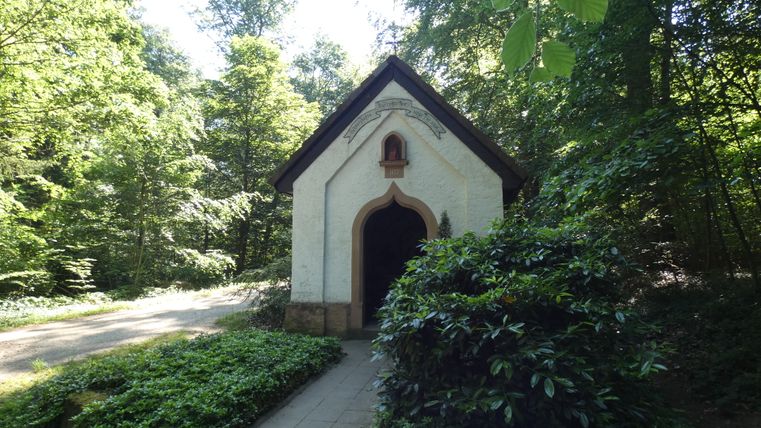 Eine kleine Kapelle im Wald mit einem Weg daneben.