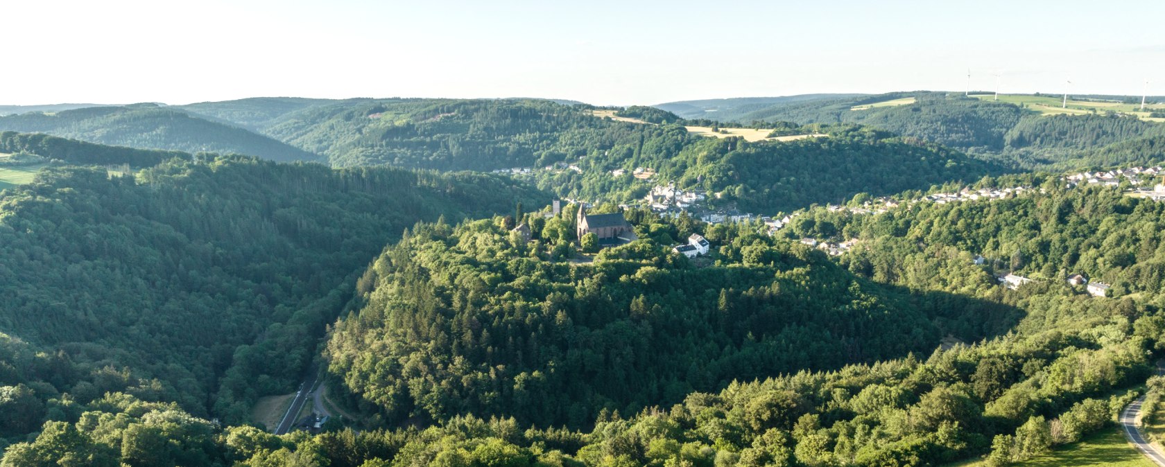 Aerial view of a green hilly landscape with a village on a hill surrounded by forests and fields under a clear sky., © Eifel Tourismus GmbH, Dominik Ketz