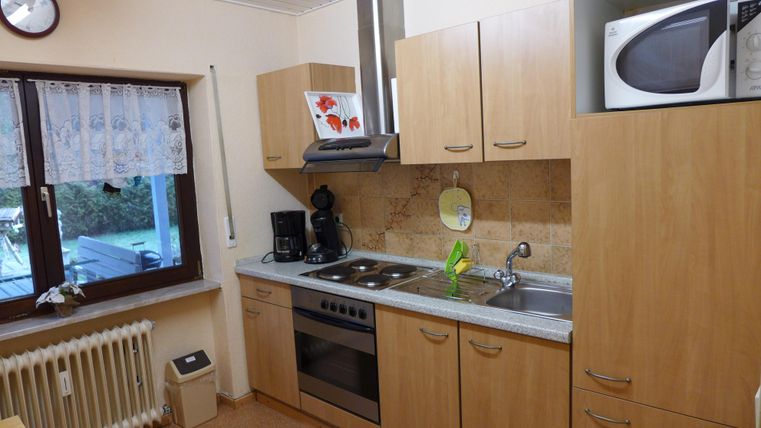 A bright kitchen with wooden furniture and modern appliances. A sheer curtain hangs at the window, and there is a cooking area as well as a sink.