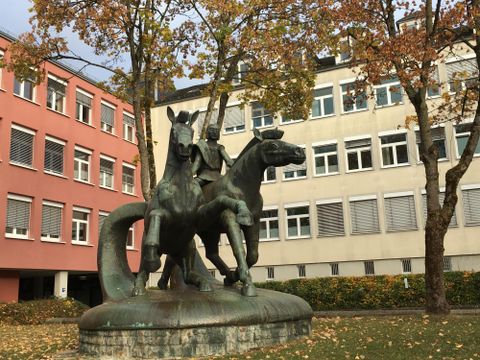 Een bronzen sculptuur van twee paarden met een ruiter bevindt zich in een stedelijk gebied. Op de achtergrond zijn moderne gebouwen en bomen met herfstbladeren te zien.