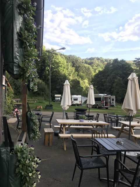 Ein einladender Außenbereich mit Tischen und Stühlen. Im Hintergrund sind grüne Hügel und Wohnmobile sichtbar.