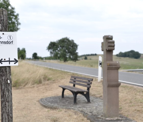 Signposts to Wißmannsdorf and a bench on a country road, surrounded by fields and trees., © Tourist-Info Bitburger Land