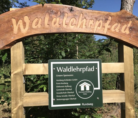 Wooden sign with the inscription 'Waldlehrpfad' and a green information board with sponsor details and a circular route sign., © TI Bitburger Land
