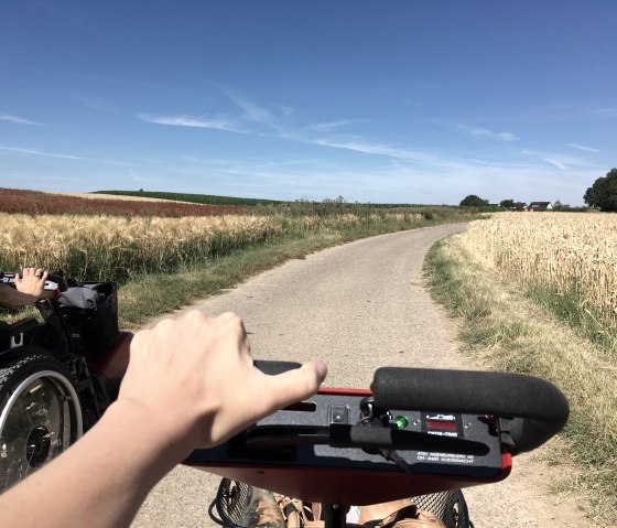 Eine Person fährt mit einem Swiss Trac auf einem ländlichen Weg, umgeben von Getreidefeldern unter blauem Himmel., © TI Bitburger Land