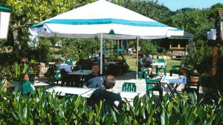 Een gezellig café in de buitenlucht met een grote parasol en groene stoelen. Omgeven door planten en bomen genieten de gasten van de ontspannen sfeer.