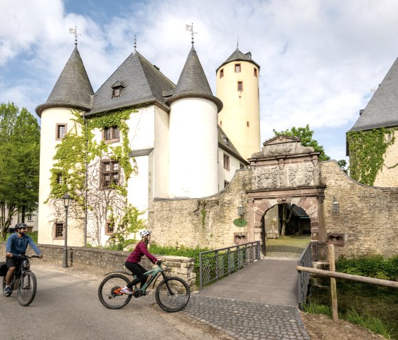 Deux cyclistes passent devant le château de Rittersdorf sur la piste cyclable de la Nims. Le château a des tours rondes et est recouvert de lierre., © Eifel Tourismus GmbH, Dominik Ketz