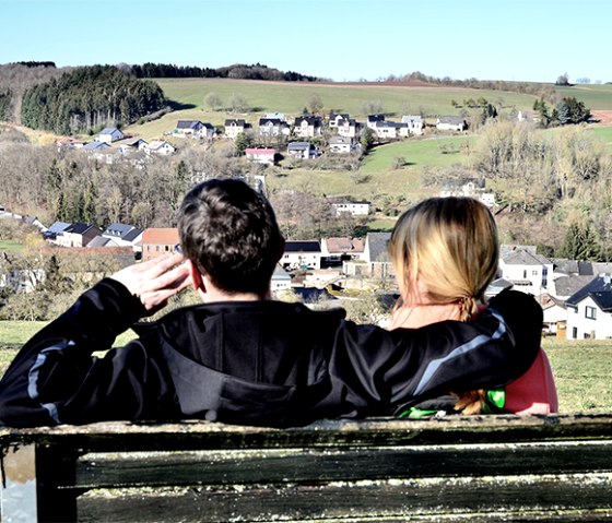 A couple sits on a bench and looks out over the village of Baustert in a green, hilly landscape., © TI Bitburger Land