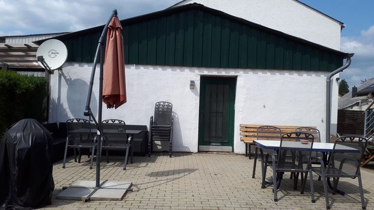 Ein gemütlicher Außenbereich mit einem Tisch und Stühlen. Im Hintergrund ist ein Haus mit einem grünen Dach und einer Markise zu sehen.