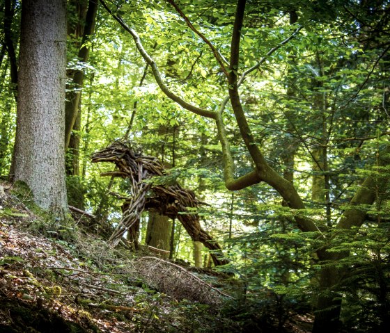 Eine Holzskulptur im Wald, die einem Tier ähnelt, umgeben von dichtem Laub und Bäumen, Teil der Kunstroute Kyllburg., © TI Bitburger Land_M.Mayer