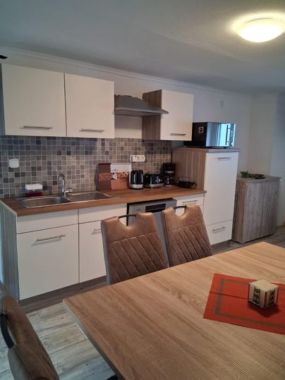 Eine moderne Küche mit hellen Möbeln und einer eleganten Holzoberfläche. Der Tisch ist mit einem roten Untersetzer dekoriert und im Hintergrund stehen verschiedene Küchengeräte.