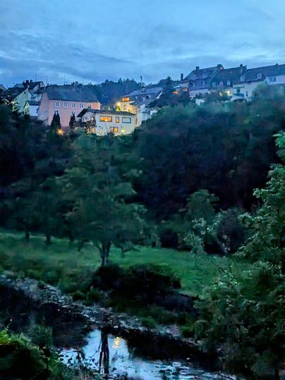 Ein ruhiges Dorf in der Dämmerung, umgeben von Bäumen und Hügeln. Ein kleiner Fluss fließt durch die Landschaft, während einige Häuser sanft beleuchtet sind.