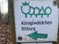 Sign with the inscription 'Königswäldchen Bitburg' and a green arrow pointing to the left. Trees and foliage can be seen in the background., © TI Bitburger Land