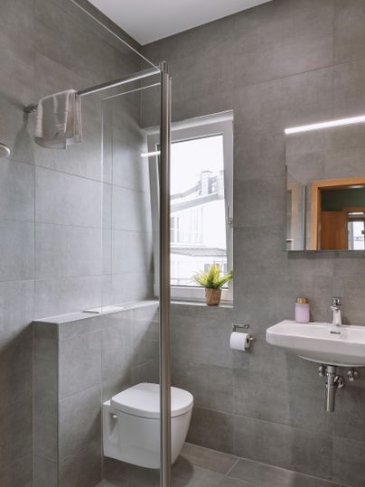 Ein modernes Badezimmer mit grauen Fliesen und einer Dusche aus Glas. Es gibt auch ein kleines Fenster und eine grüne Pflanze, die frischen Akzent setzt.