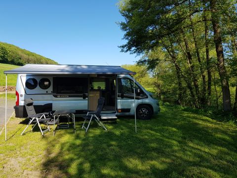 Een camper staat op een weiland, omgeven door bomen. Twee opklapstoelen zijn voor het voertuig geplaatst, ideaal om te ontspannen in de natuur.