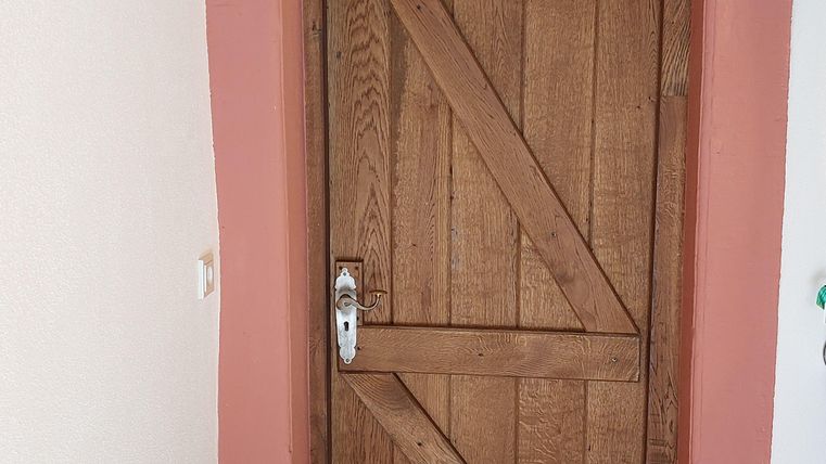 Een houtkleurige deur met een diagonaal patroon in een roze frame. De omgeving heeft een lichte muur en een betegelde vloer.