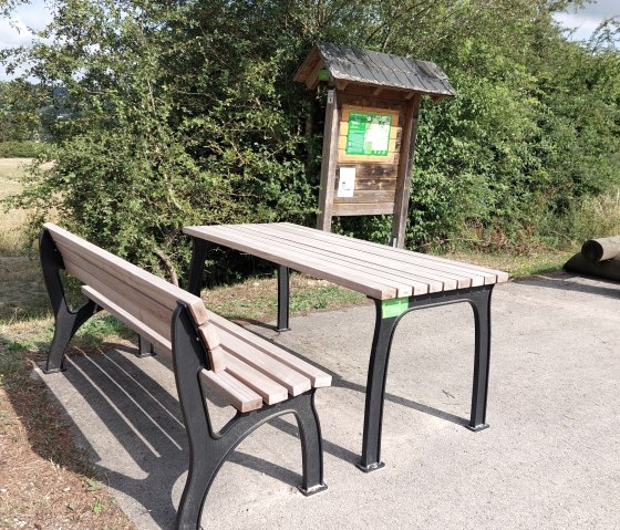 Een picknicktafel met een bankje staat naast een houten informatiebord op een geplaveid plein omringd door struiken en bomen., © Otmar Schröder