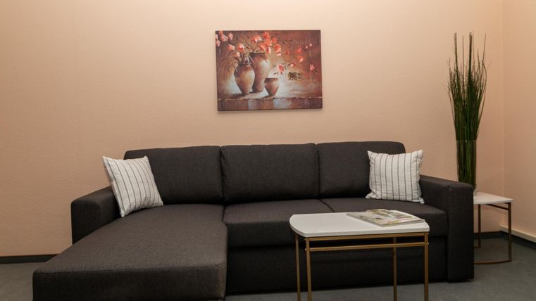 Ein modernes Wohnzimmer mit einem grauen L-förmigen Sofa und Kissen. An der Wand hängt ein Gemälde mit Vasen und Blumen, daneben steht ein grüner Pflanzenständer.