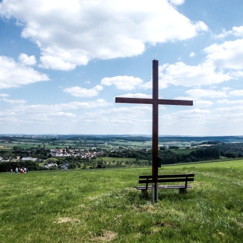Houten kruis op een weiland met een bankje, op de achtergrond een weids landschap en een dorp onder een blauwe lucht met wolken., © TI Bitburger Land