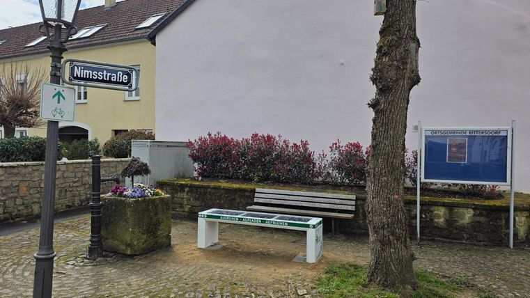 A quiet corner with a bench and a street name sign "Nimstraße". In the background, flowers and a tree are visible, surrounded by paved ground.