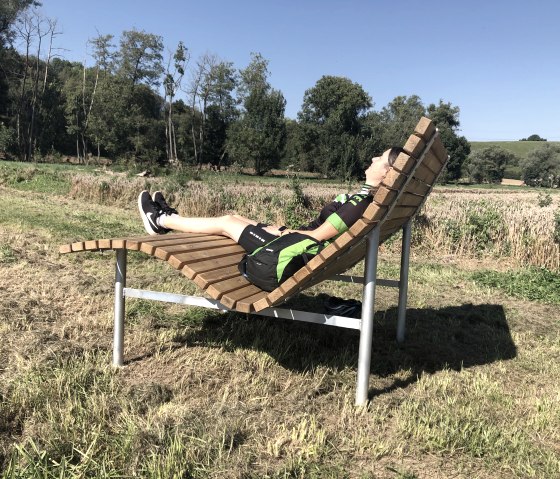 Een persoon ontspant op een gebogen houten bank in een groen landschap onder een blauwe hemel., © Tourist Information Bitburger Land