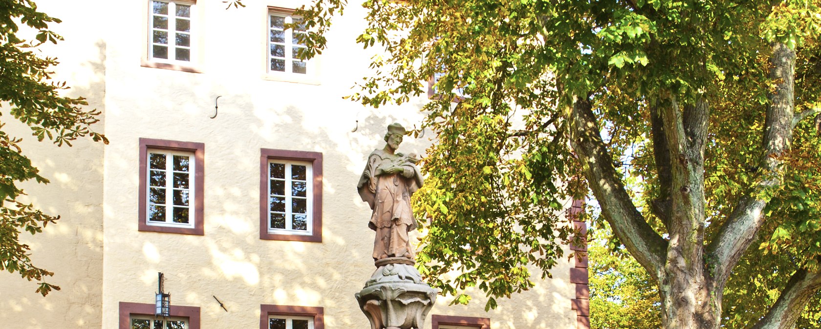 Eine Statue vor einem historischen Gebäude in Dudeldorf, umgeben von Bäumen und einer Steinmauer. Ein Schild weist auf den "Dahmsbrunnen" hin., © TI Bitburger Land