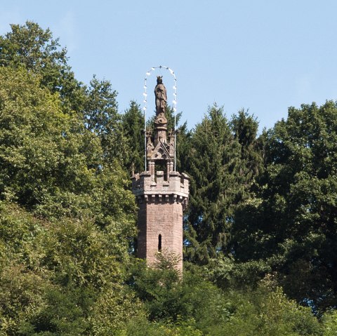 De Mariazuil in Kyllburg rijst op uit het dichte bos. Een standbeeld staat op een stenen toren, omringd door groene bomen en een blauwe lucht., © TI Bitburger Land