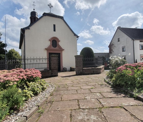 Dorfkirche in Sefferweich, umgeben von blühenden Beeten und einem gepflasterten Weg, unter einem blauen Himmel mit Wolken., © Tourist-Information Bitburger Land