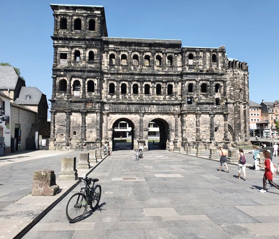 De Porta Nigra in Trier bij zonnig weer. Mensen wandelen, een fiets op de voorgrond. Historische architectuur tegen een blauwe lucht., © TI Bitburger Land, Steffi Wagner