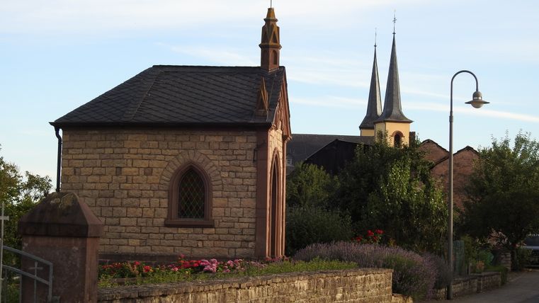 Kleine Kapelle aus Stein mit spitzem Dach, umgeben von Blumen und Bäumen, im Hintergrund Kirchtürme.