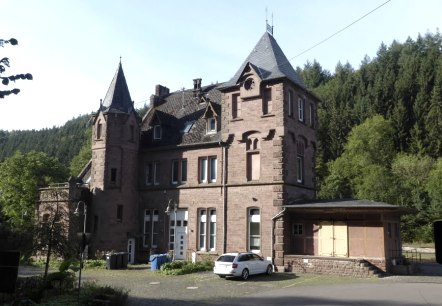 Historisches Bahnhofsgebäude aus rotem Backstein mit Türmen, umgeben von Bäumen und einem geparkten Auto., © Eifelverein Ortsgruppe Speicher