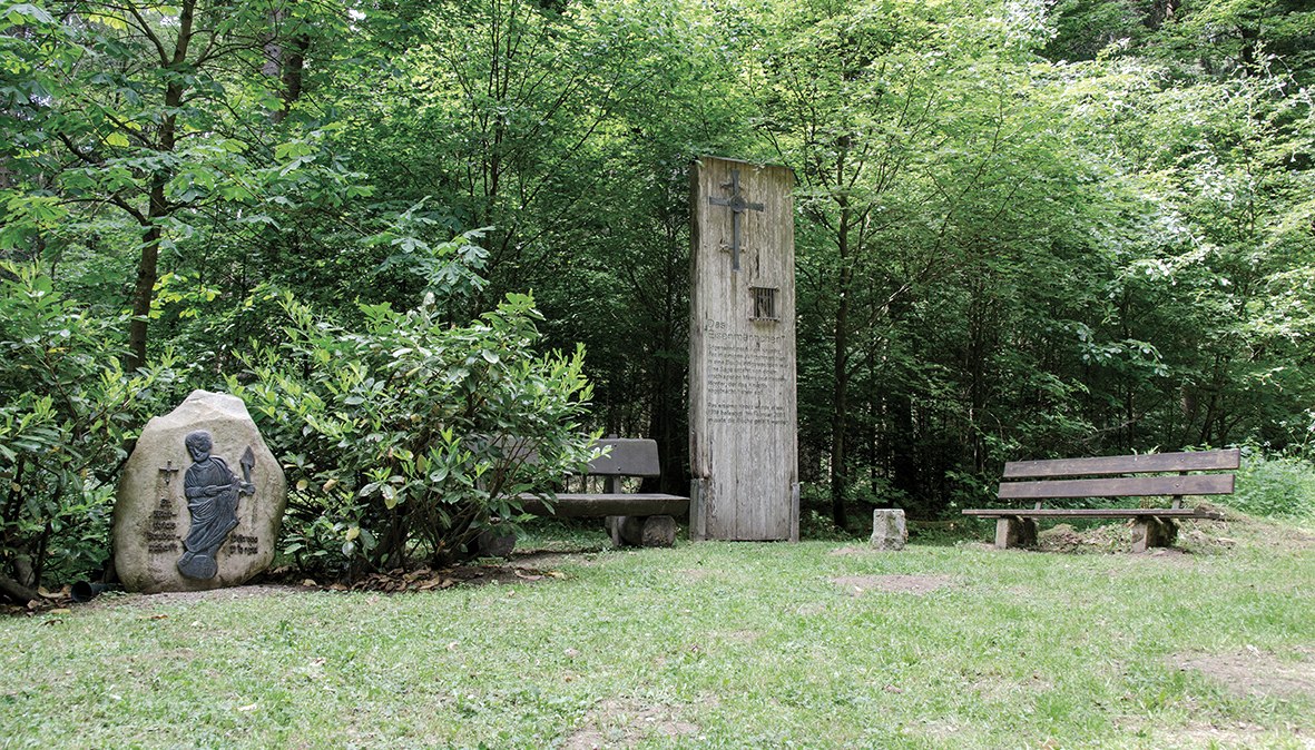 A wooden monument and a stone relief in the forest, surrounded by benches and trees. The scene is calm and inviting., &copy; TI Bitburger Land