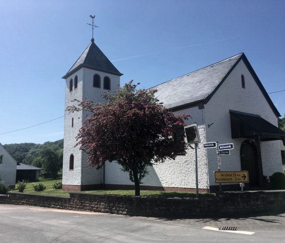 Weiße Kirche mit Turm und rotem Baum im Vordergrund, blauer Himmel. Straßenschilder weisen nach Arzfeld und Koosbüsch., © Fellmann