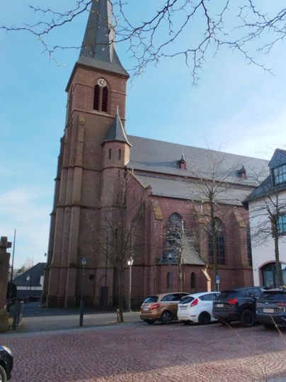 An old church with a tall tower stands in the foreground. In the background, some vehicles and a clear blue sky can be seen.