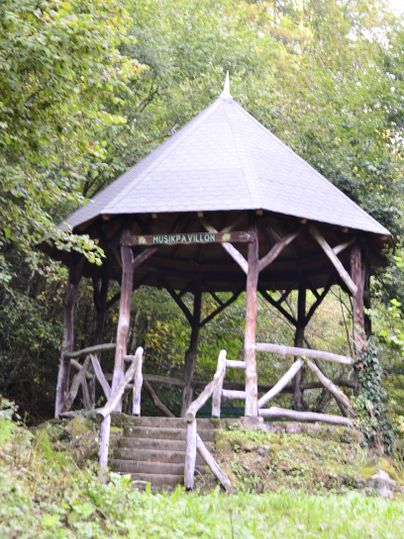 Ein hölzerner Musikpavillon im Wald mit einem spitzen Dach und einer Treppe, die hinaufführt.