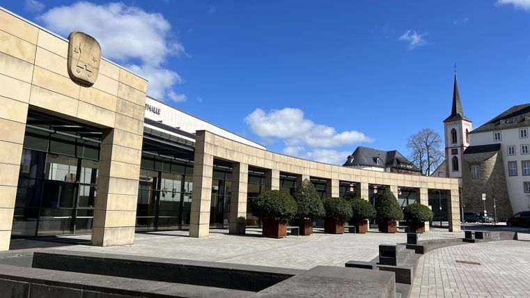 Een modern gebouw met glazen gevel en planten voor de ingang. Op de achtergrond is een kerktoren en een heldere blauwe lucht te zien.