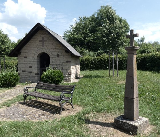 Eine kleine Steinkapelle mit Kreuz, daneben eine Bank und ein steinernes Kreuz in einer grünen, ländlichen Umgebung., © TI Bitburger Land