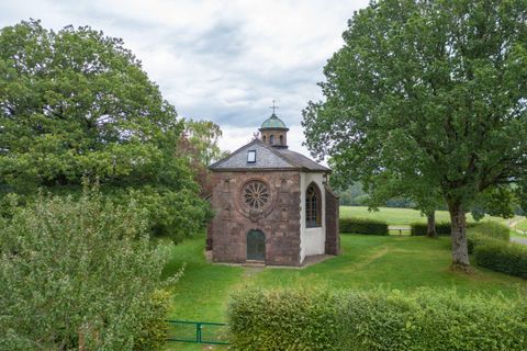 Eine kleine Kapelle steht inmitten von grünen Bäumen. Der Himmel ist bewölkt, und die Umgebung ist friedlich.